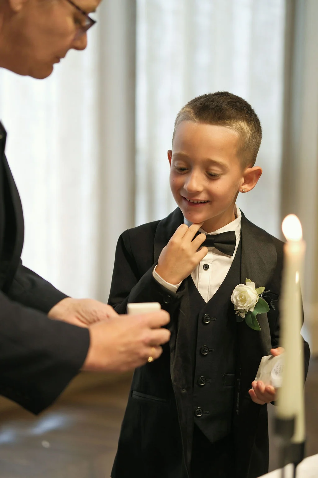 A young page boy in a smart black tuxedo and bow tie smiles during a wedding ceremony.