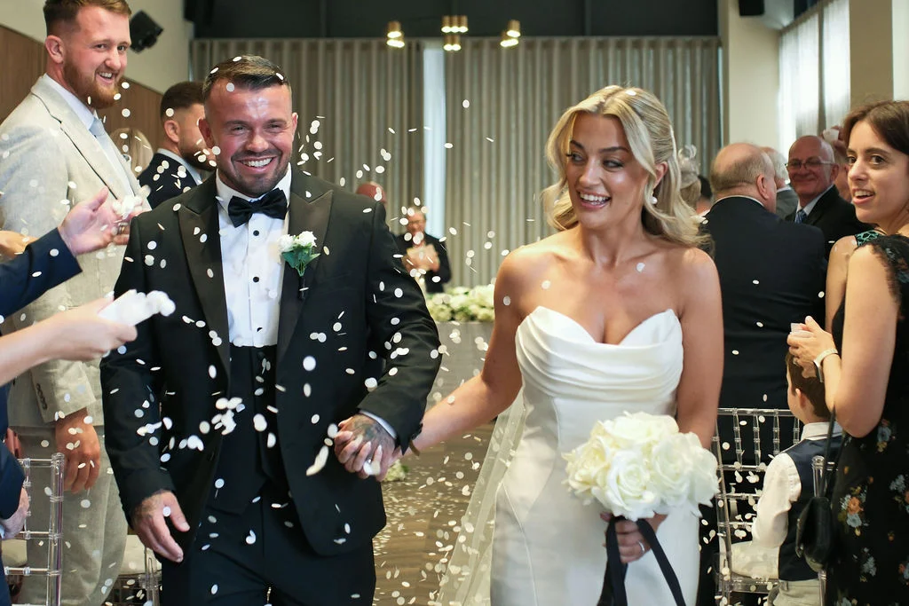 A joyous newlywed couple walk hand in hand down the aisle as white confetti falls around them, with the smiling groom in a black tuxedo and the radiant bride in a strapless white gown carrying a bouquet of white roses.