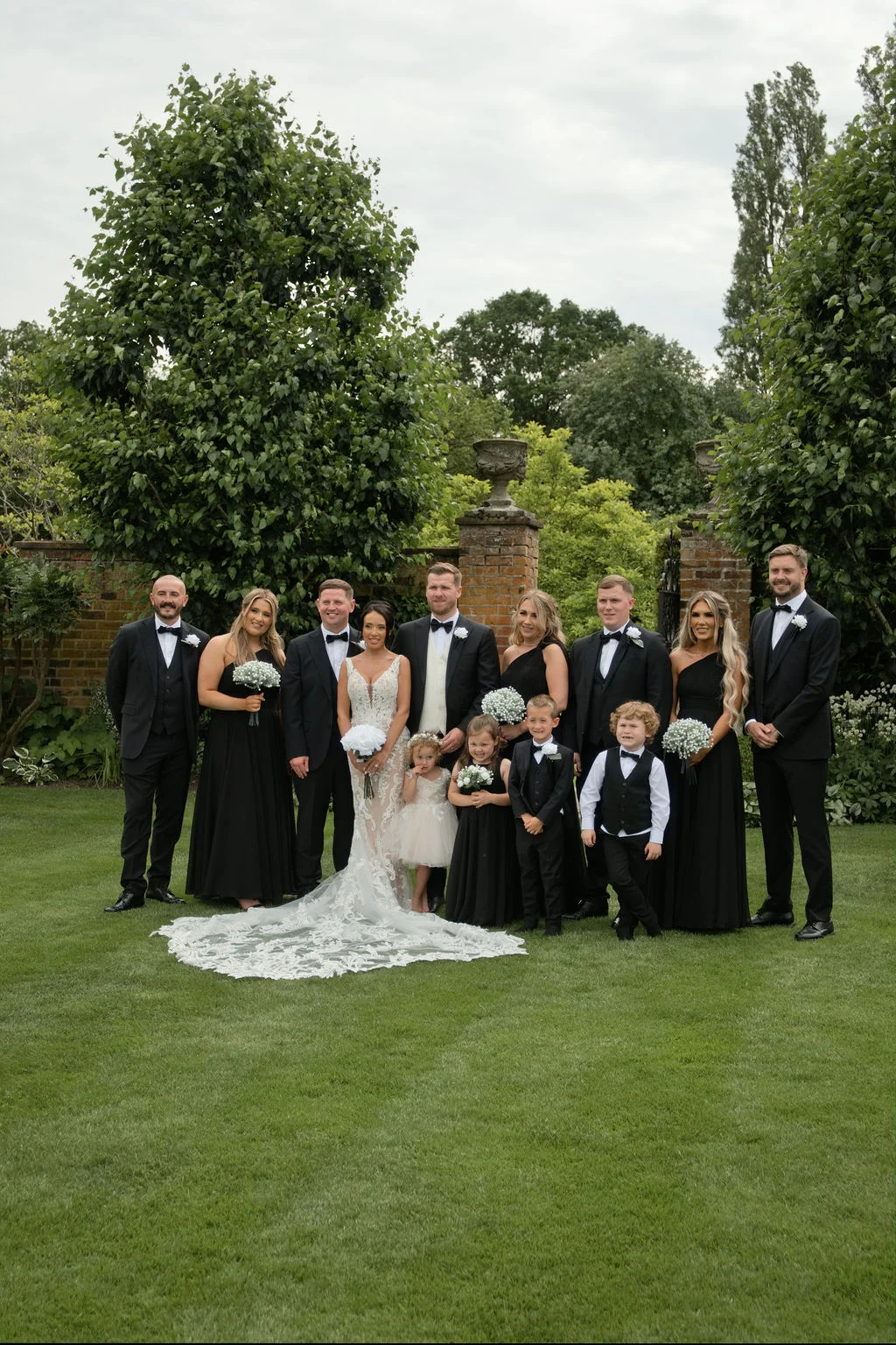 A full-length group portrait of the bride, groom, groomsmen in black tuxedos, bridesmaids in matching black dresses, and children standing together on a lush green lawn.
