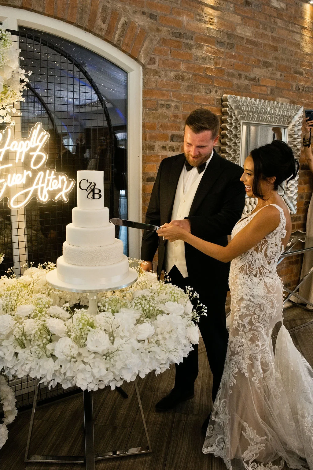 A smiling bride and groom cut a four-tiered white wedding cake surrounded by an abundant display of white hydrangeas against an exposed brick wall.