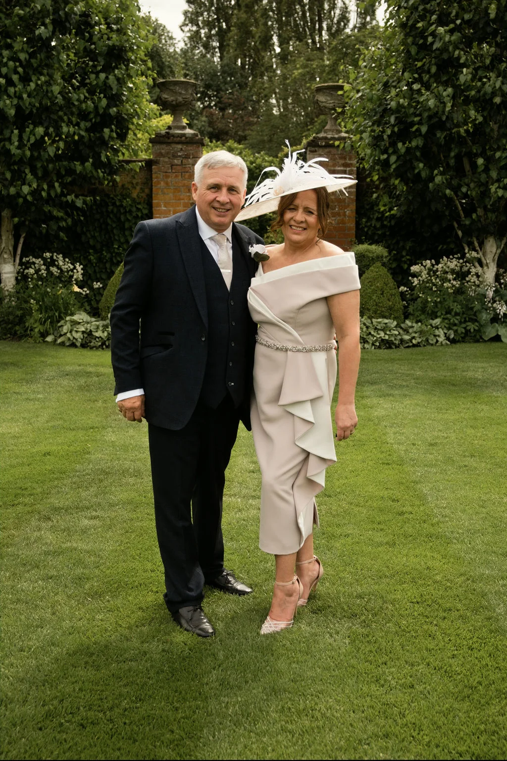 A smiling couple standing on a manicured lawn outdoors, with the woman wearing an elegant taupe dress and feathered fascinator and the man in a smart navy three-piece suit.