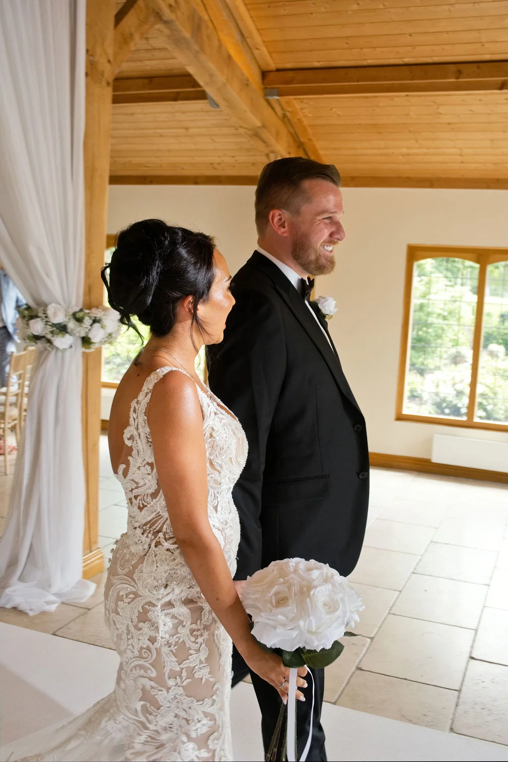 A bride in a lace mermaid gown and a groom in a black tuxedo stand side by side during an indoor ceremony inside a light-filled oak framed barn.