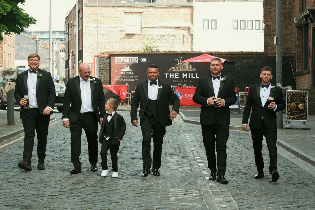 The groom and his groomsmen stride together down a historic cobblestone city street wearing classic black tuxedos and bow ties.