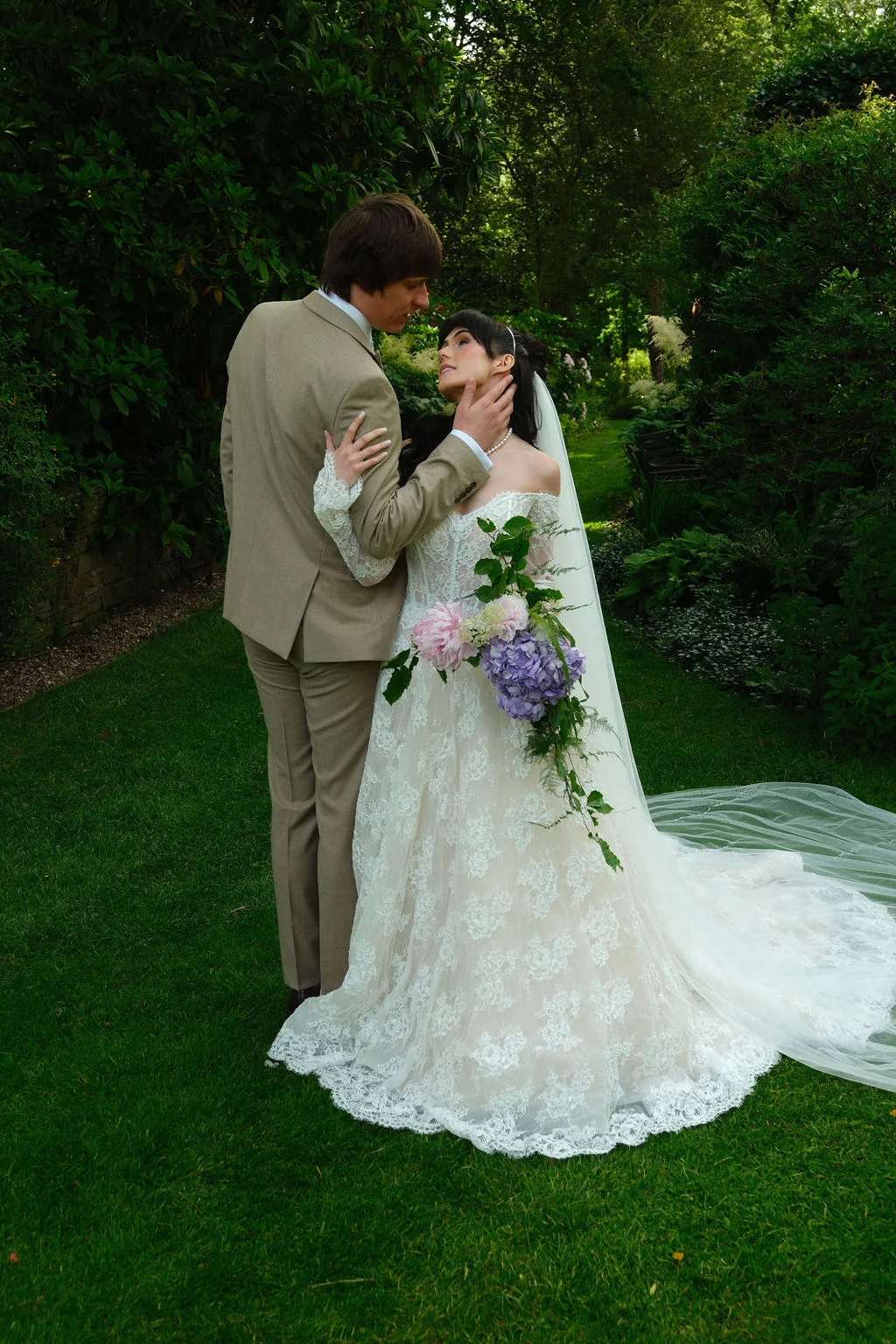 A newlywed couple embraces in a lush garden setting, with the bride looking up lovingly while holding a cascading floral bouquet.
