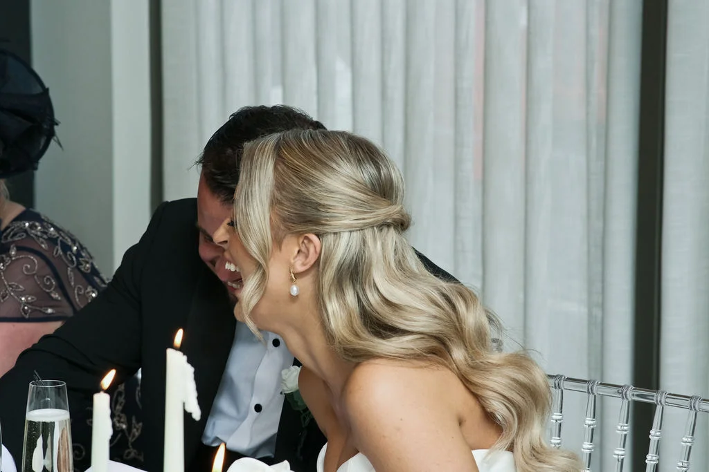 A newlywed couple shares a joyful laughing moment together at the top table during their wedding reception, illuminated by candlelight.