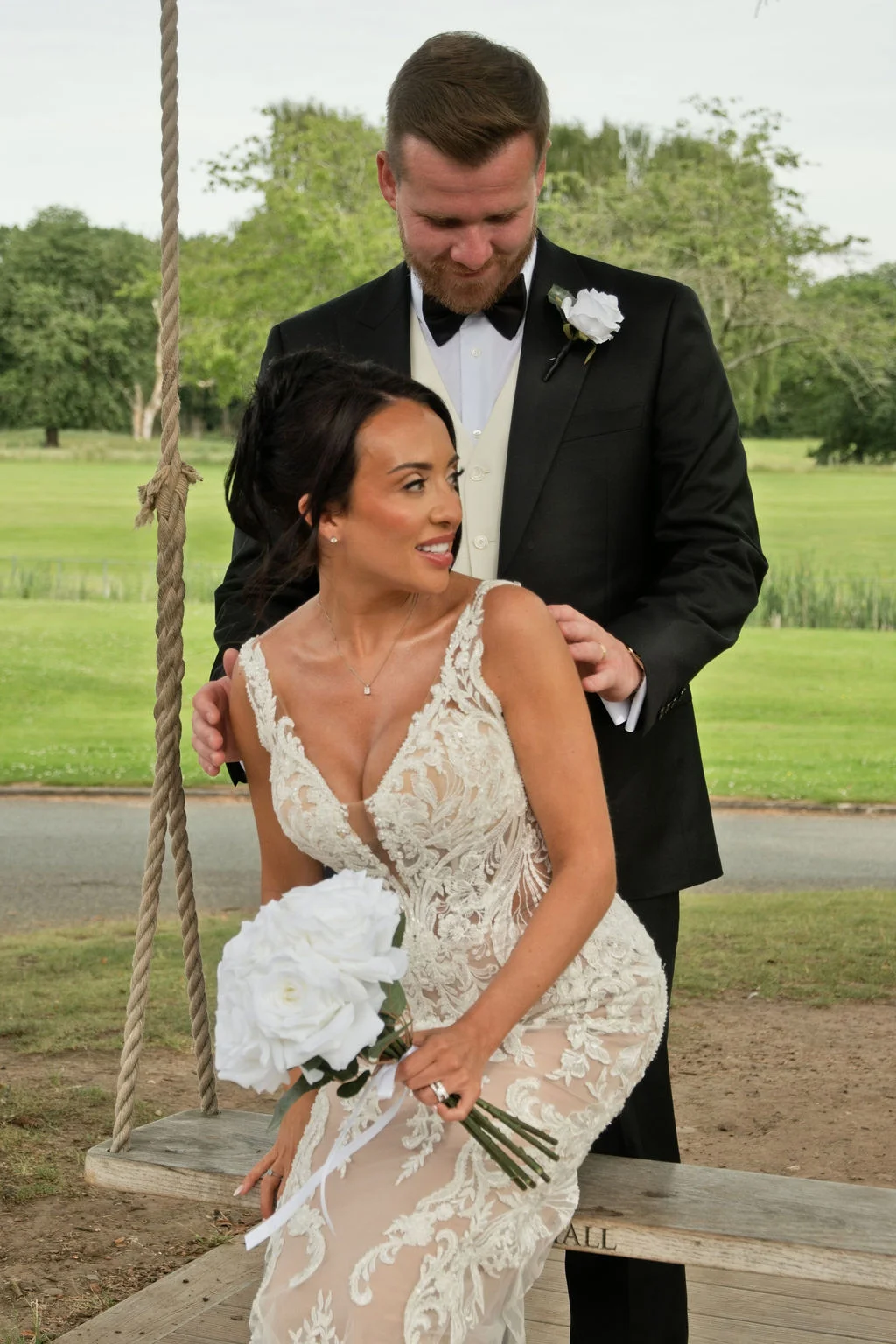 A newlywed couple poses outdoors by a rustic wooden swing, with the bride holding a bouquet of white roses while wearing a lace gown.