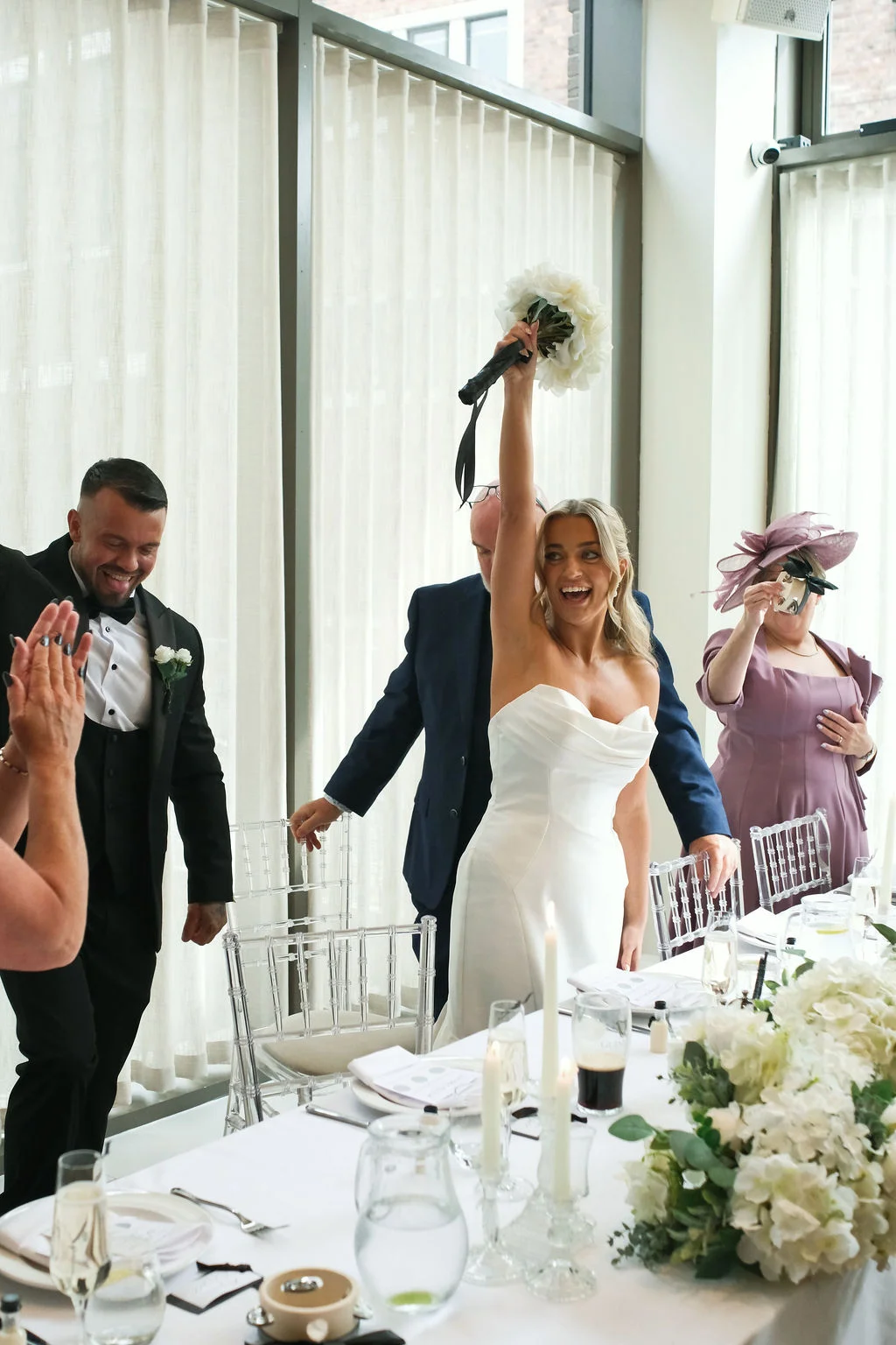 A joyful bride raises her bouquet triumphantly in the air while standing at the top table next to a laughing groom in a black tuxedo.