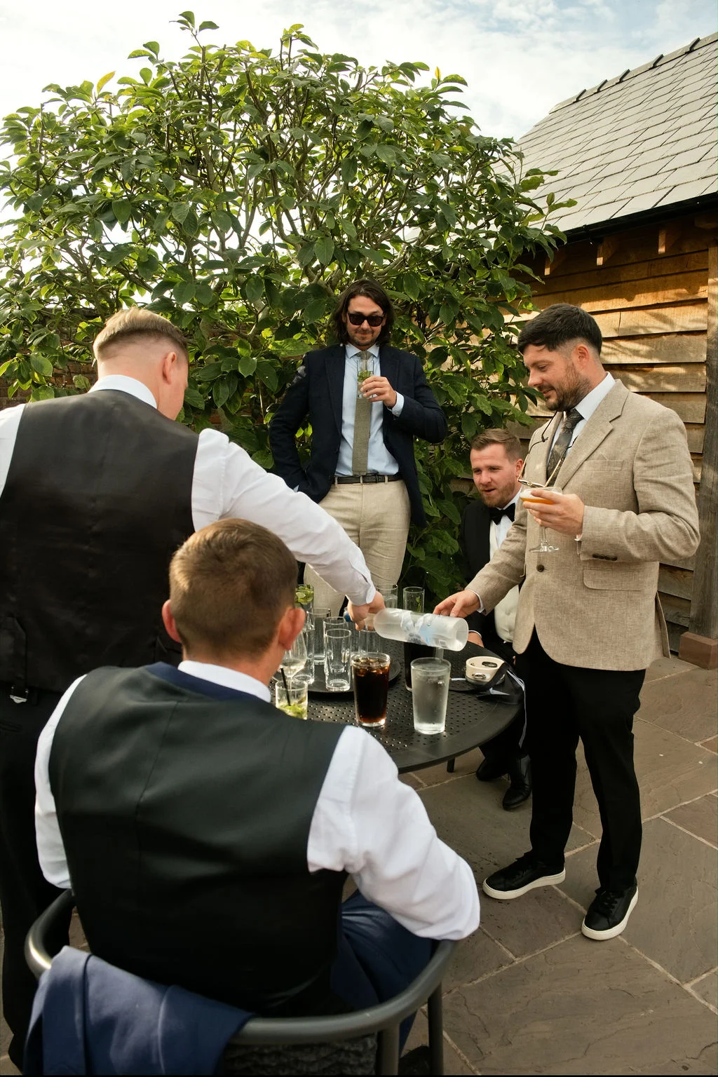 A group of groomsmen and wedding guests gather outdoors around a table during the drinks reception, with one guest pouring drinks in the sunlight.