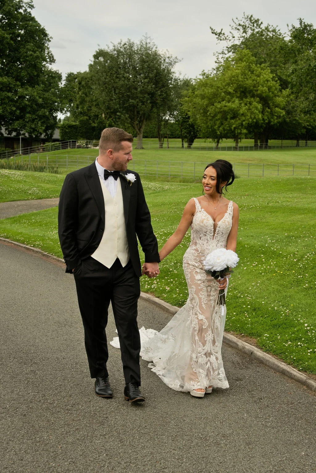 A newlywed couple holds hands and smiles at each other while walking along a paved path bordered by lush green grass and trees.
