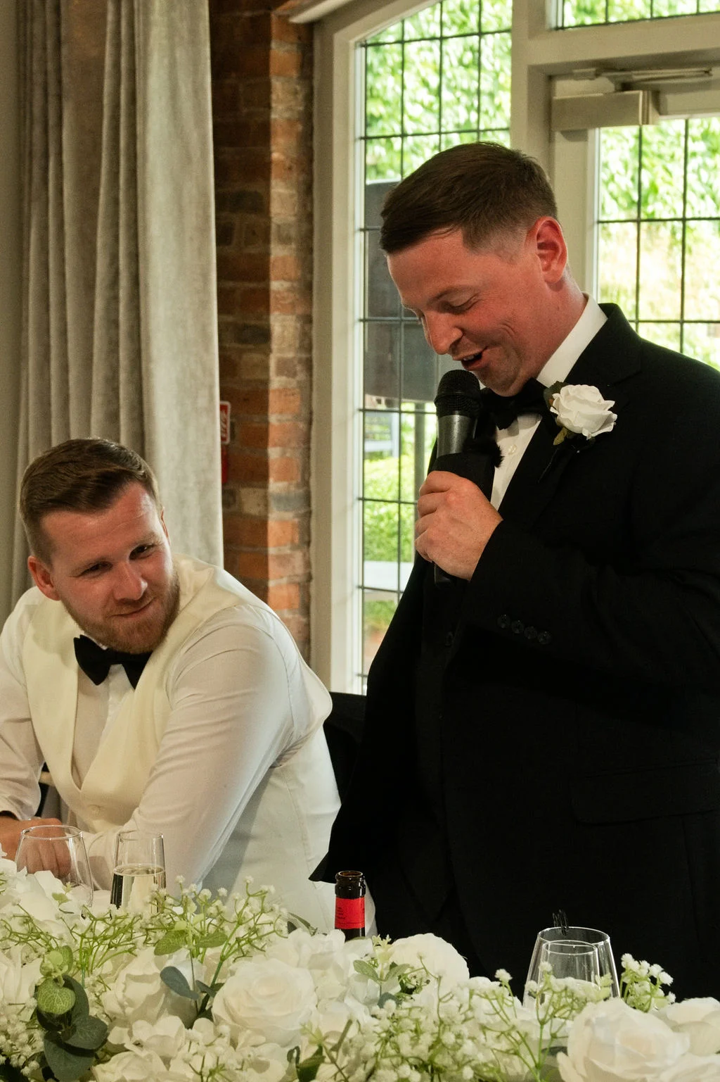 A groom in a black tuxedo stands delivering a wedding speech with a microphone, while another man seated nearby smiles during the reception.