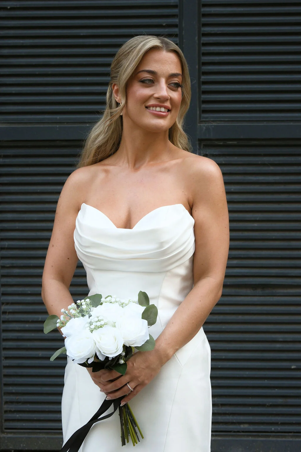 A glowing bride poses with a hand-tied bouquet of white roses and eucalyptus against a dark slatted backdrop, wearing a strapless ruched gown and half-up hair.