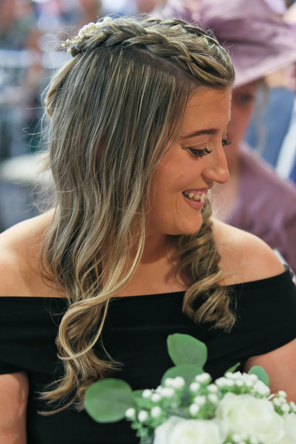 A smiling bride wearing an off-the-shoulder dress with a half-up braided hairstyle adorned with pearls, holding a white rose and eucalyptus bouquet.