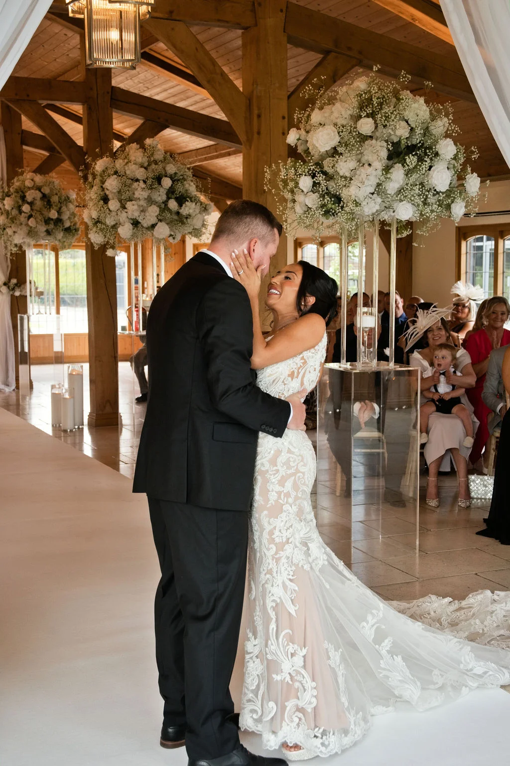 A newlywed couple share an intimate first kiss during their ceremony inside a rustic oak-framed barn decorated with towering white floral arrangements.