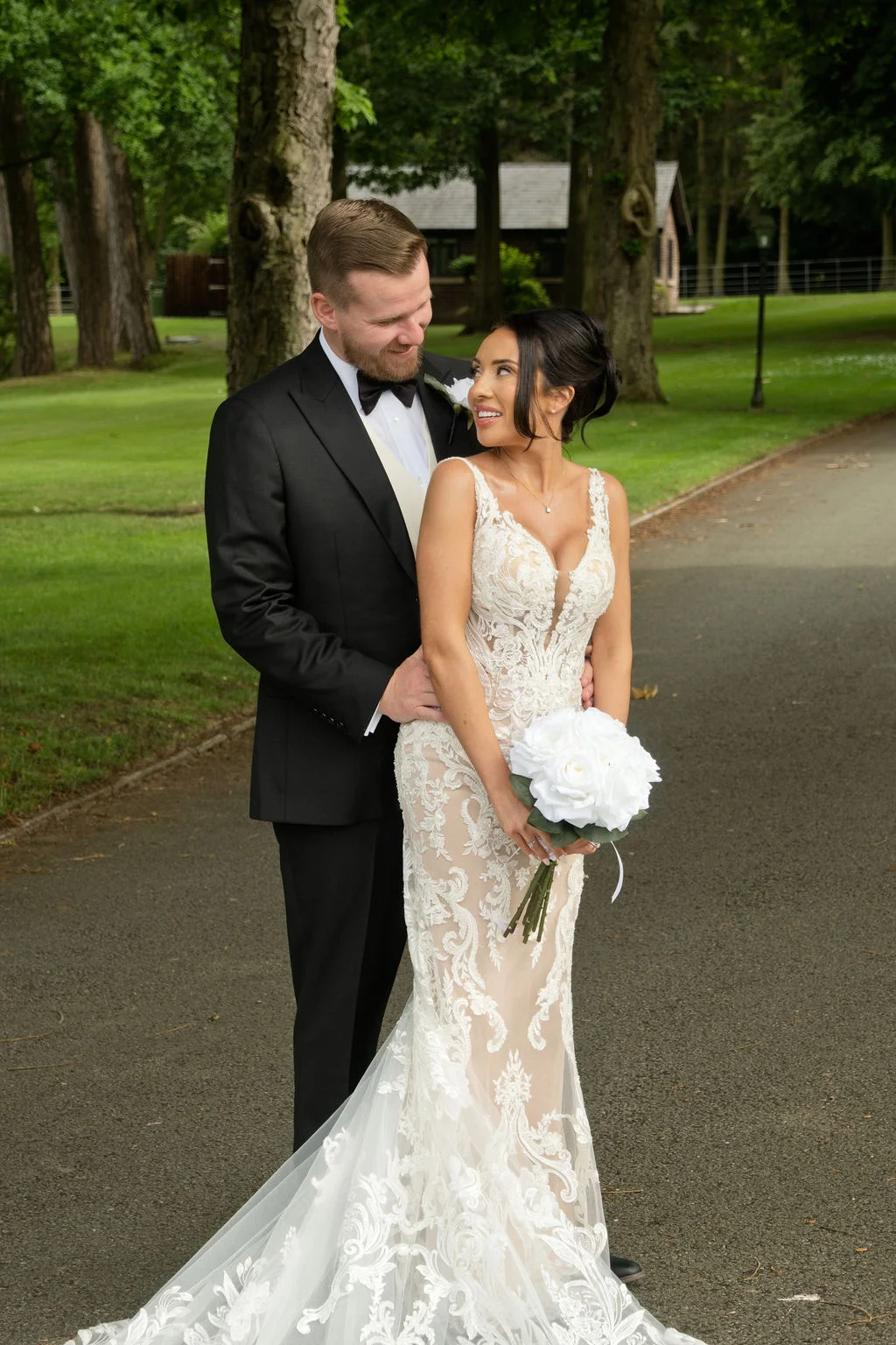 A newlywed couple shares a loving glance outdoors on a paved pathway flanked by lush green lawns and tall trees.