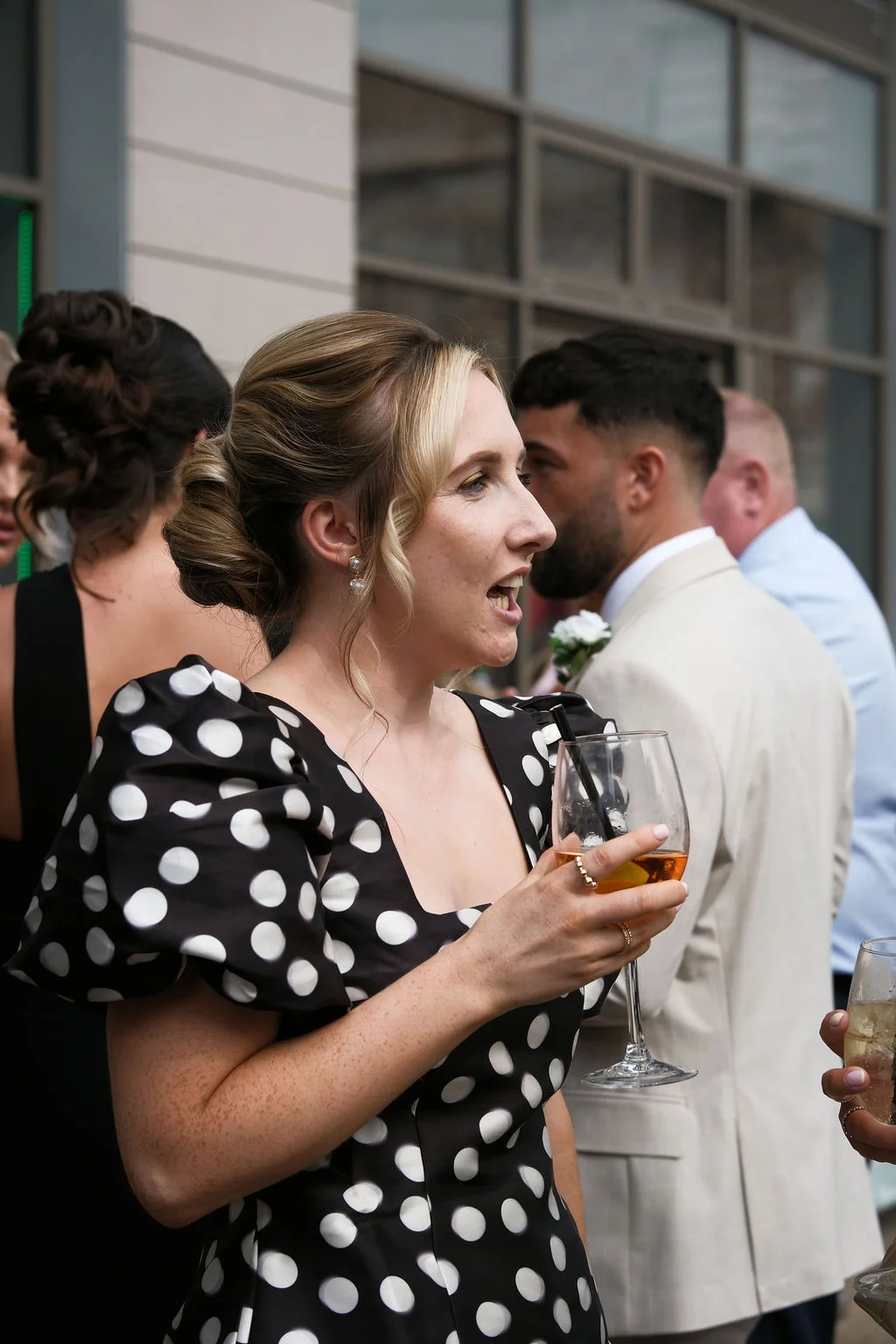 A wedding guest wearing a black and white polka dot dress with puff sleeves holds a wine glass while mingling outdoors during the drinks reception.