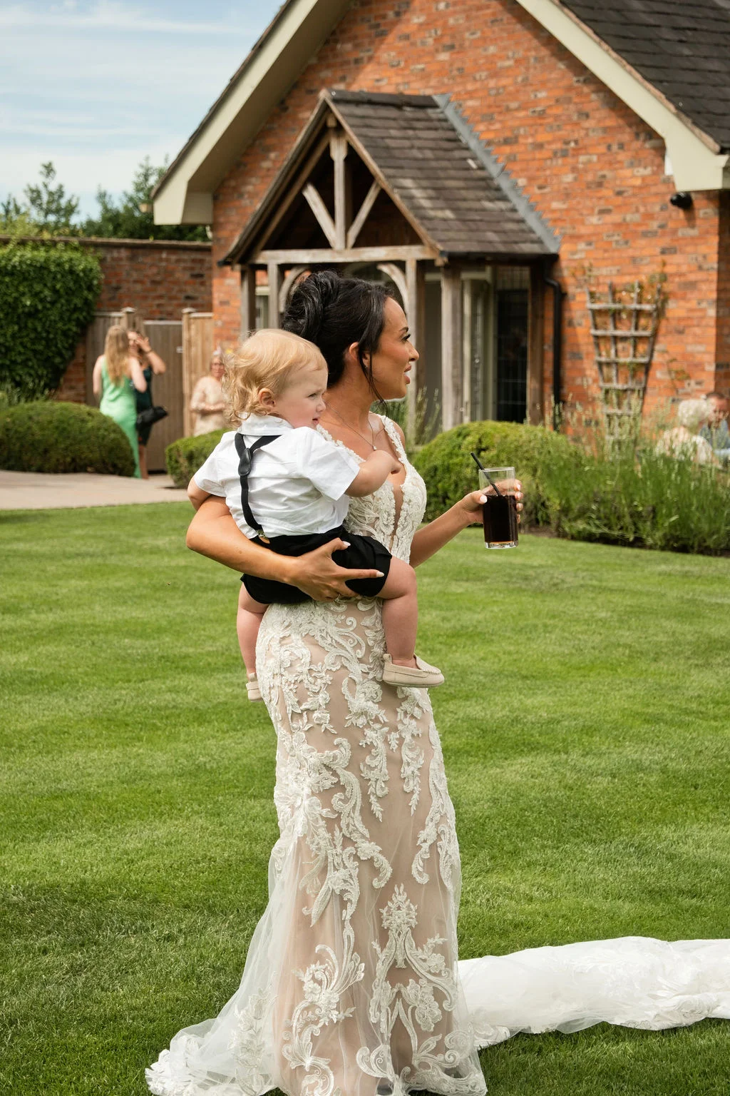 A bride in a detailed lace mermaid dress walks across a lush green lawn holding a young page boy in a white shirt and braces, while holding a dark drink during a summer drinks reception.