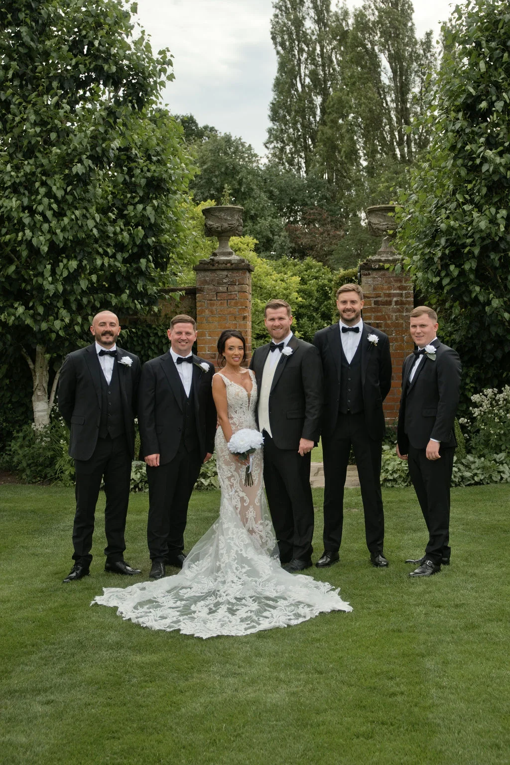 The bride in a stunning lace gown with a dramatic train stands centrally, surrounded by the groom and four groomsmen in classic black tuxedos in a manicured garden setting.
