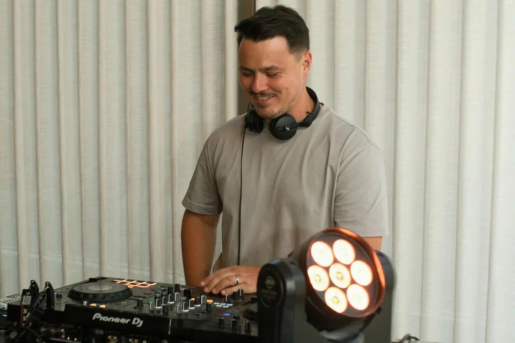 A wedding DJ with headphones around his neck smiles down while operating a Pioneer DJ mixer in front of white curtains.