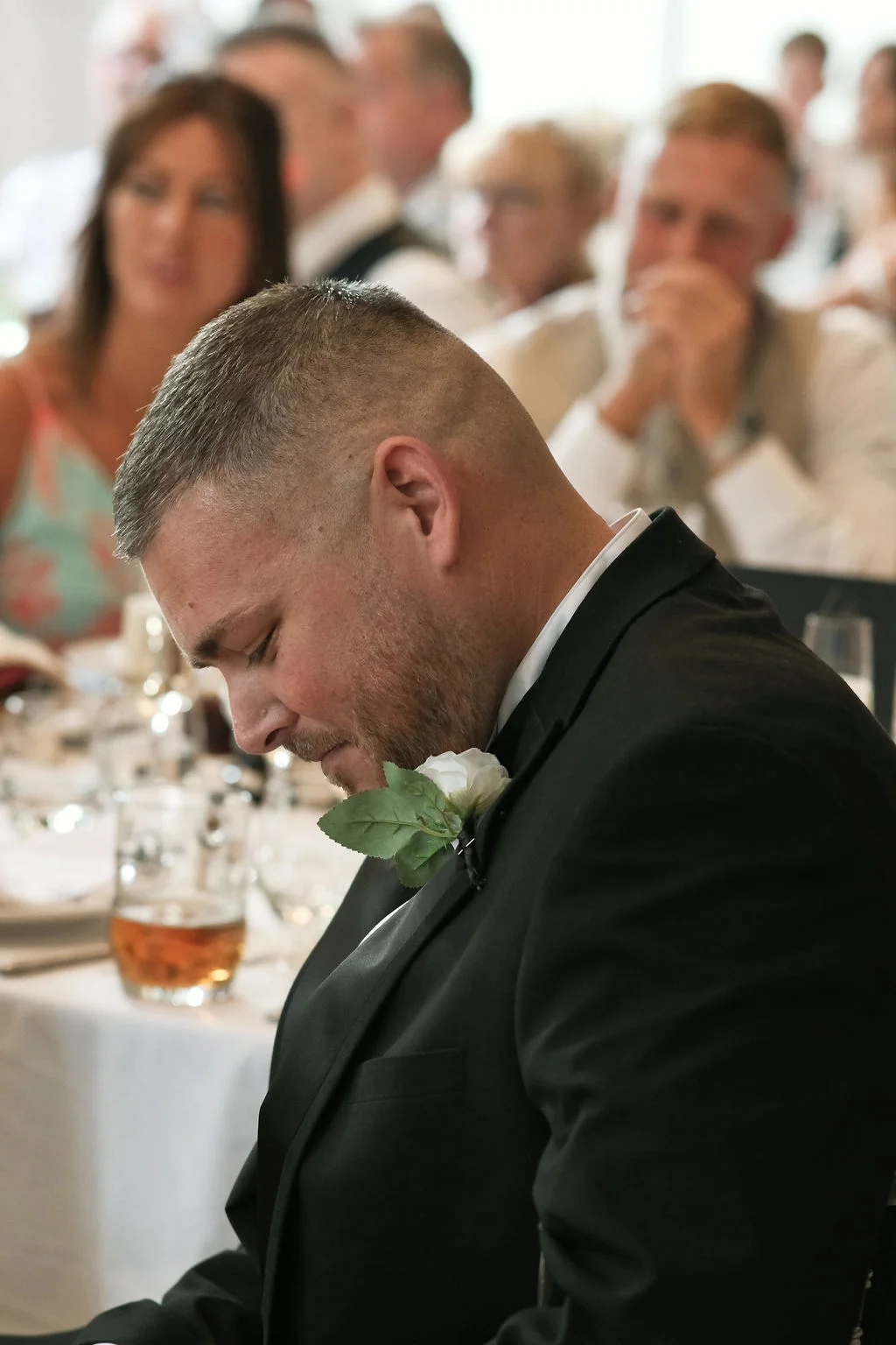 A groom wearing a black tuxedo with a white rose buttonhole sits with his head bowed during the wedding speeches as guests look on in the background.