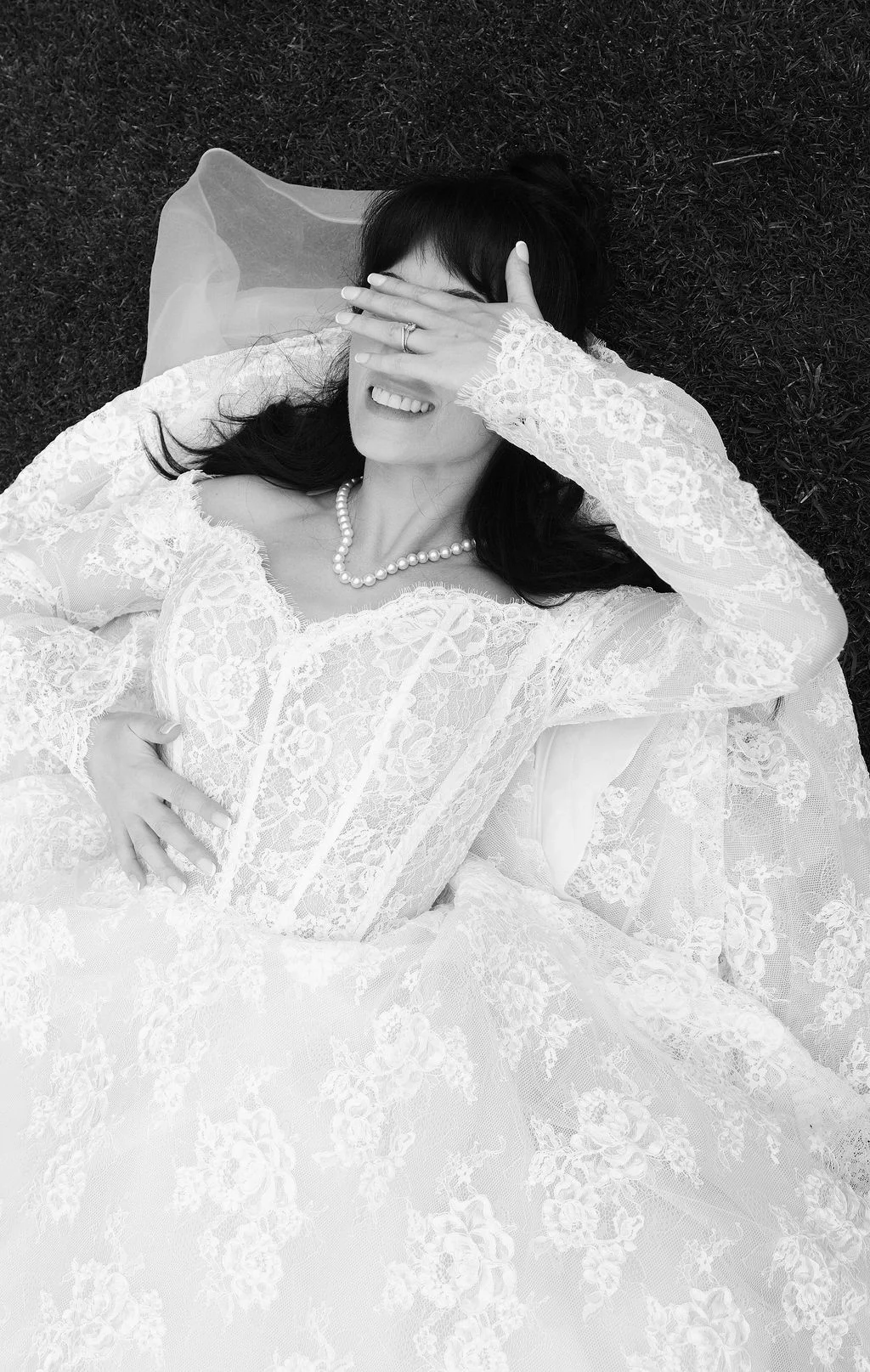 A joyful bride wearing a lace off-the-shoulder gown and pearl necklace lies back on the grass, laughing with her hand over her face.