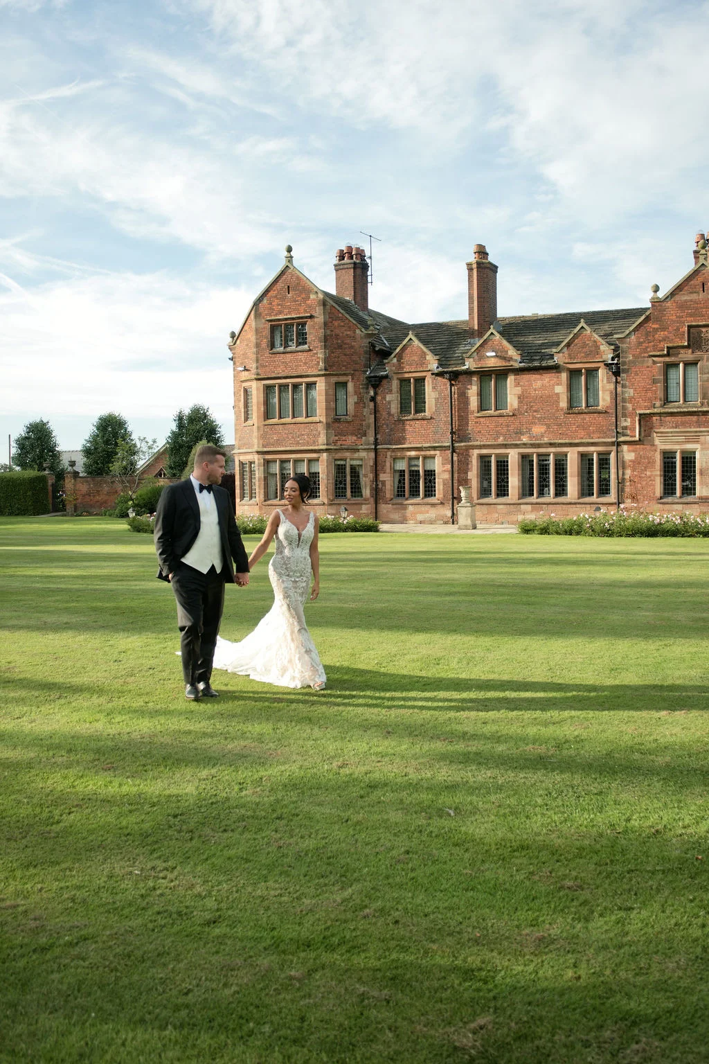 A newlywed couple walks hand in hand across a vast green lawn in front of a grand red brick manor house under a bright sky.