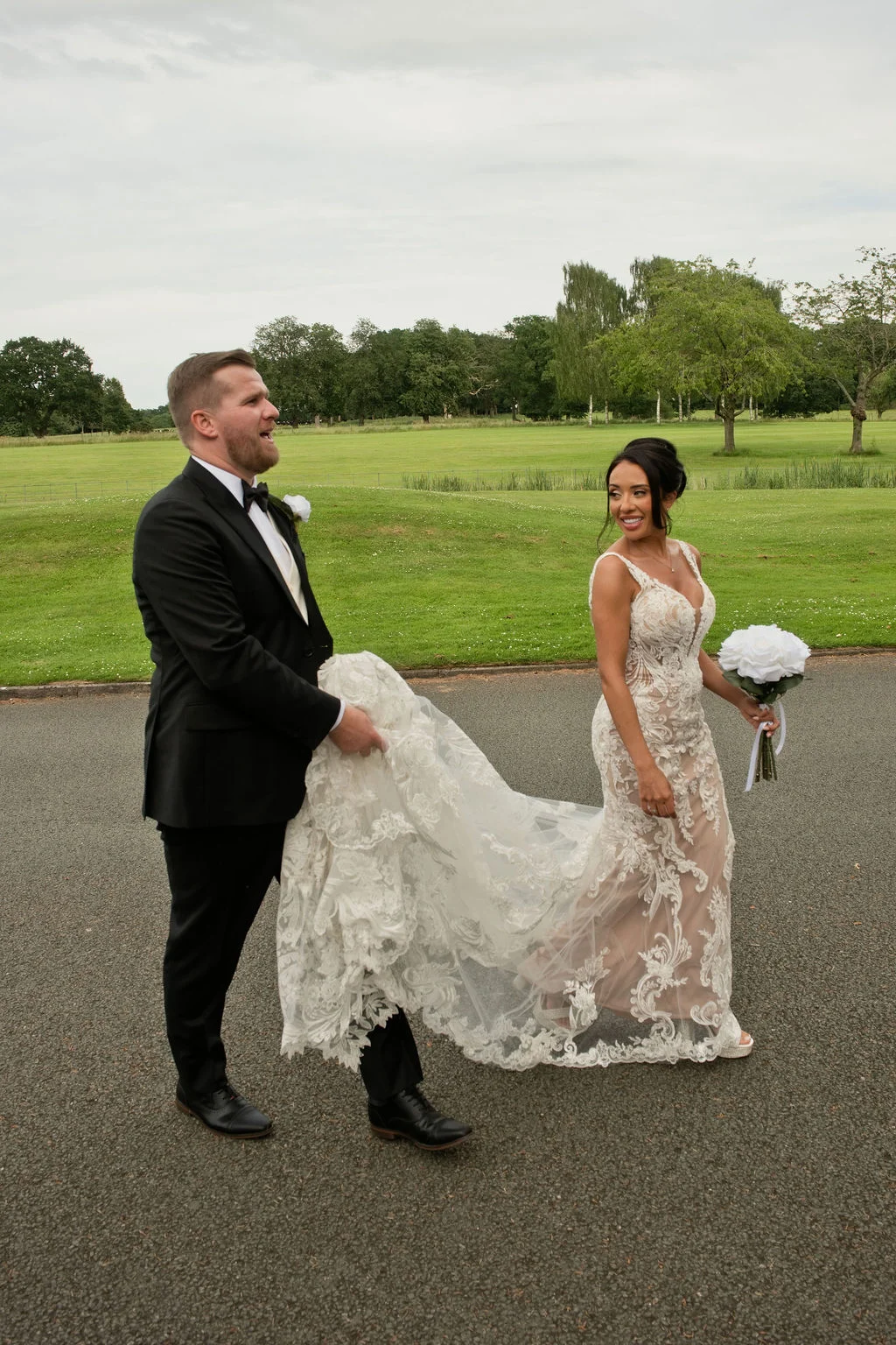 A newly married couple walk together along a path, with the smiling groom in a black tuxedo carrying the bride's long lace train while she looks back at him holding a white bouquet.