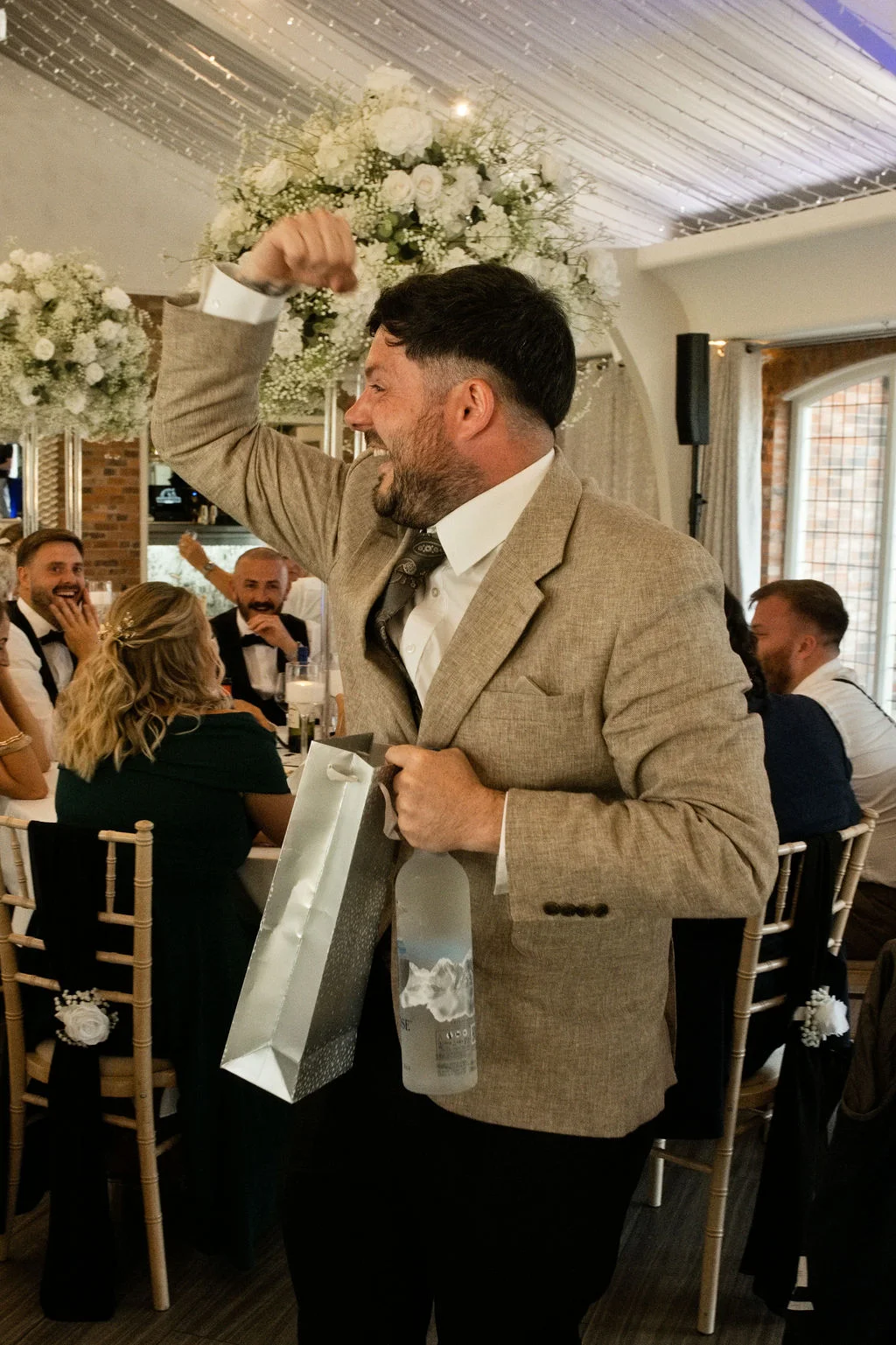 A joyful groom in a beige suit reacts excitedly during the wedding speeches, holding a bottle of vodka and a gift bag while guests laugh at the tables.