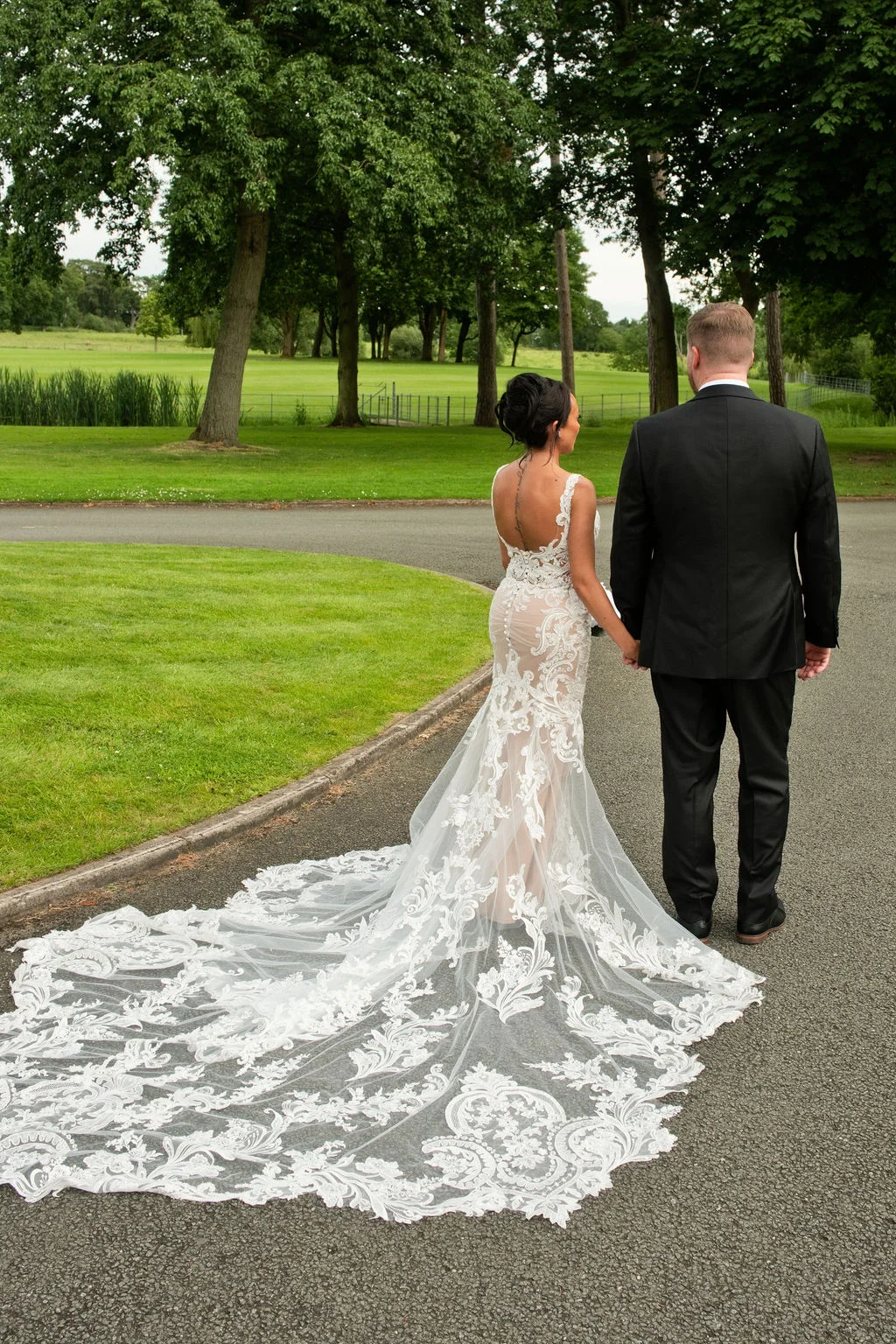 A newlywed couple walking hand in hand along a pathway in a lush green park, showcasing the bride's dramatic lace gown with a long train.