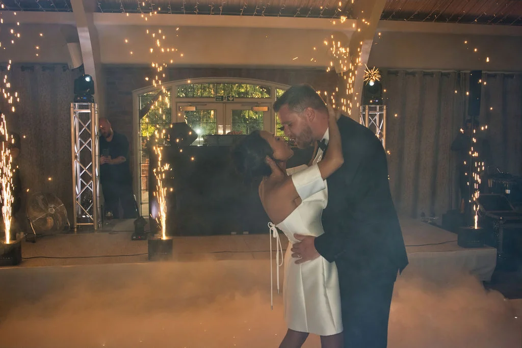 A newlywed couple share a dip kiss during their first dance on a smoke-filled dance floor surrounded by dramatic cold sparks.