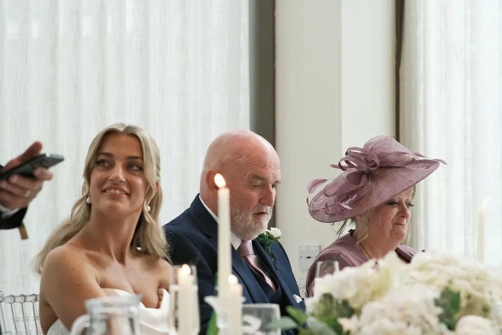 Wedding guests seated at the top table listen intently and smile during the wedding reception speeches, with lit candles and floral arrangements in the foreground.