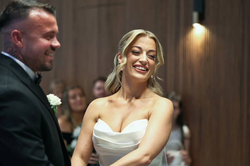 A smiling bride laughing happily during the wedding ceremony next to the groom in a modern wood panelled venue.