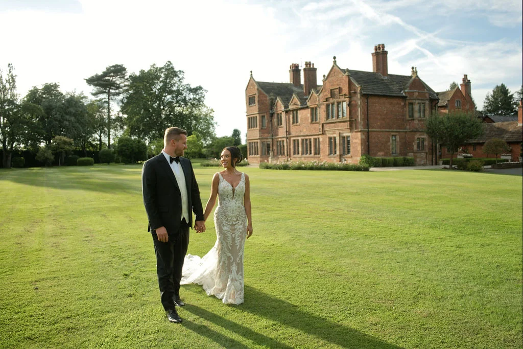 A bride and groom walk hand in hand across a manicured lawn in front of a grand manor house during golden hour.