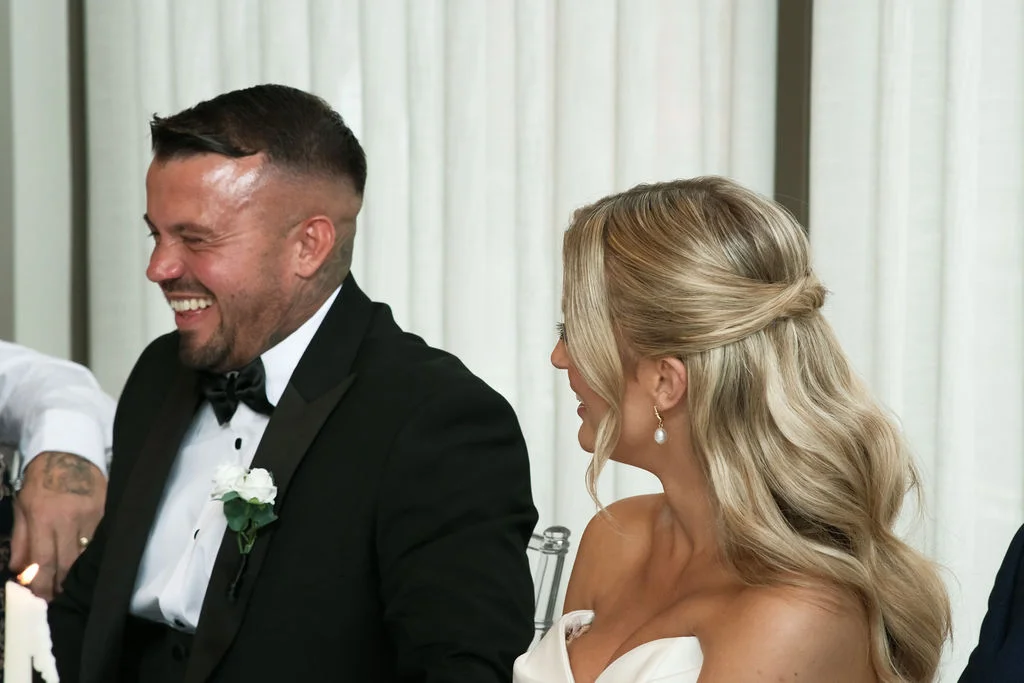 A laughing groom in a black tuxedo with a bow tie and buttonhole sits next to his smiling bride with half-up wavy blonde hair during the wedding reception speeches.