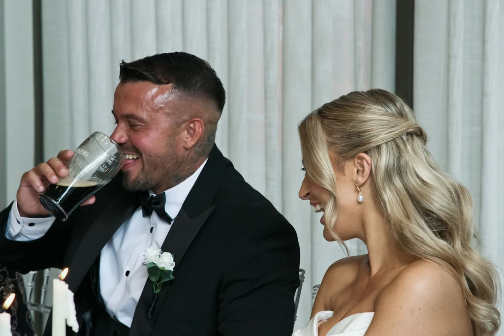 A smiling groom drinks from a pint glass while seated next to his laughing bride at the wedding reception table.