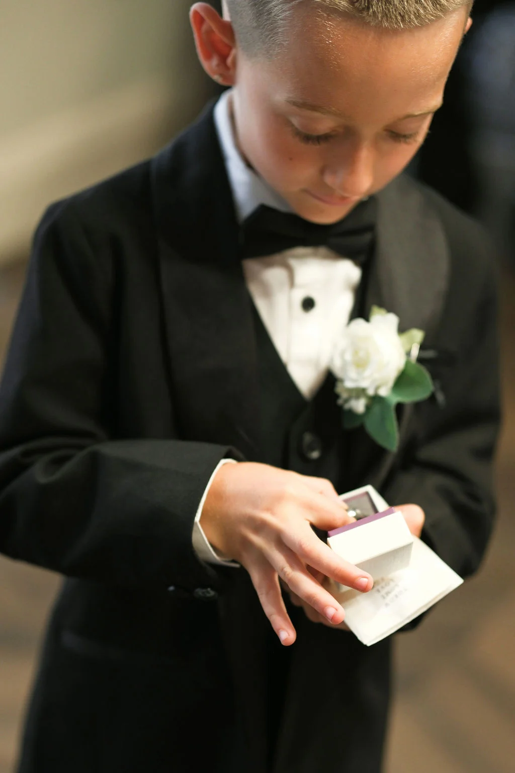 A young page boy dressed in a sharp black tuxedo and bow tie opens a small ring box while carefully holding it during the wedding ceremony.