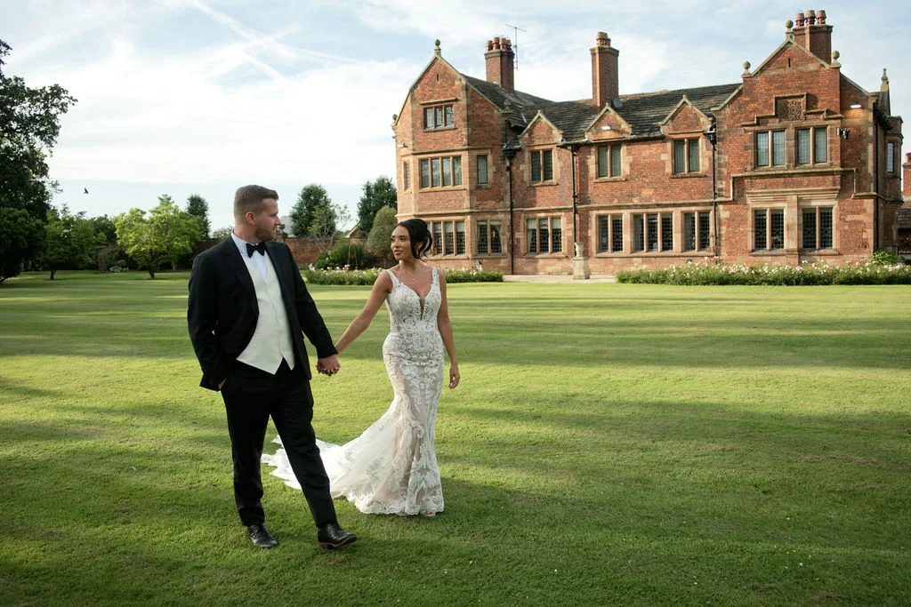 A bride and groom walk hand in hand across a manicured lawn in front of a grand red brick country house.