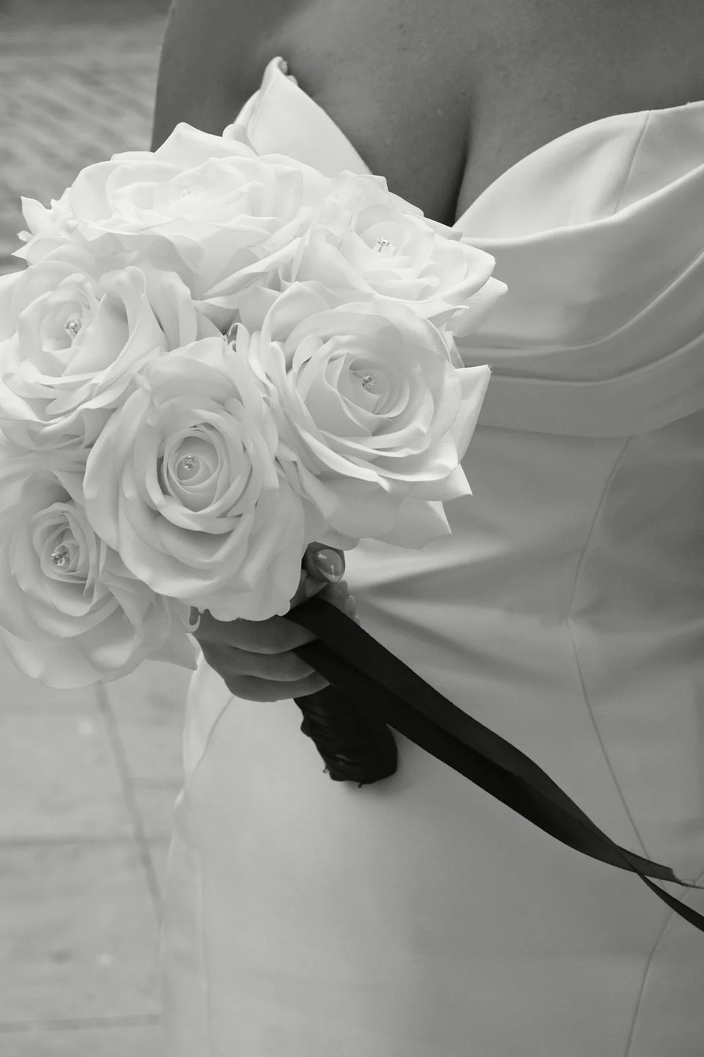 A bride in a satin gown with a sweetheart neckline holds a round hand-tied bouquet of white roses in this monochrome photograph.