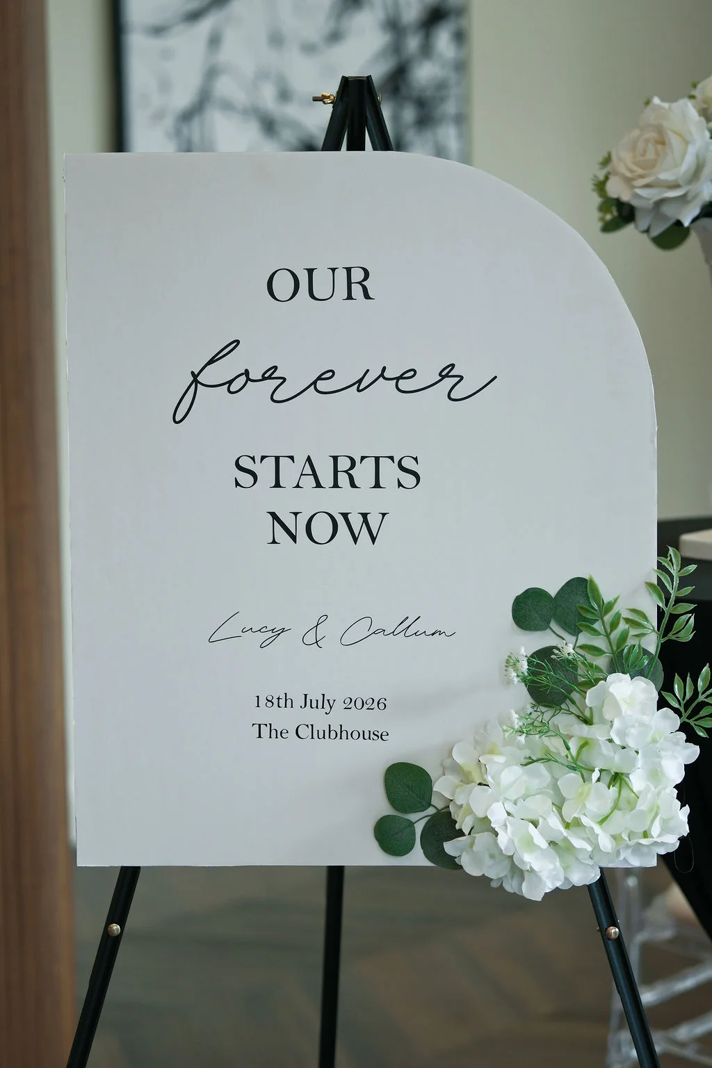 A modern arched white wedding welcome sign featuring black lettering and a floral arrangement of white roses and eucalyptus on a black easel.