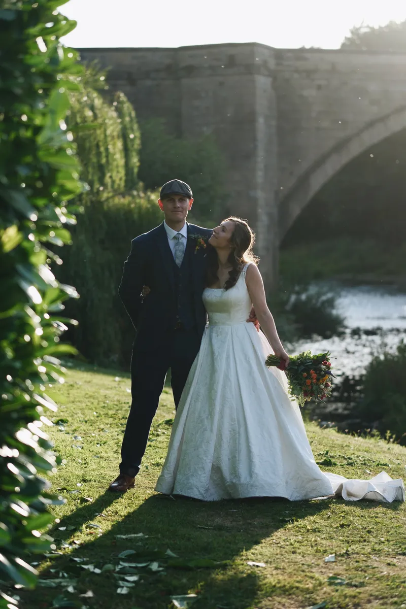 A newlywed couple poses together by a river with a stone bridge in the background during golden hour, with the bride looking up lovingly at the groom.