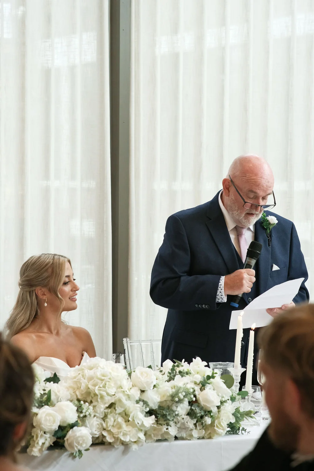 The father of the bride delivers a heartfelt speech holding a microphone while the smiling bride listens attentively beside a lush white floral centrepiece.