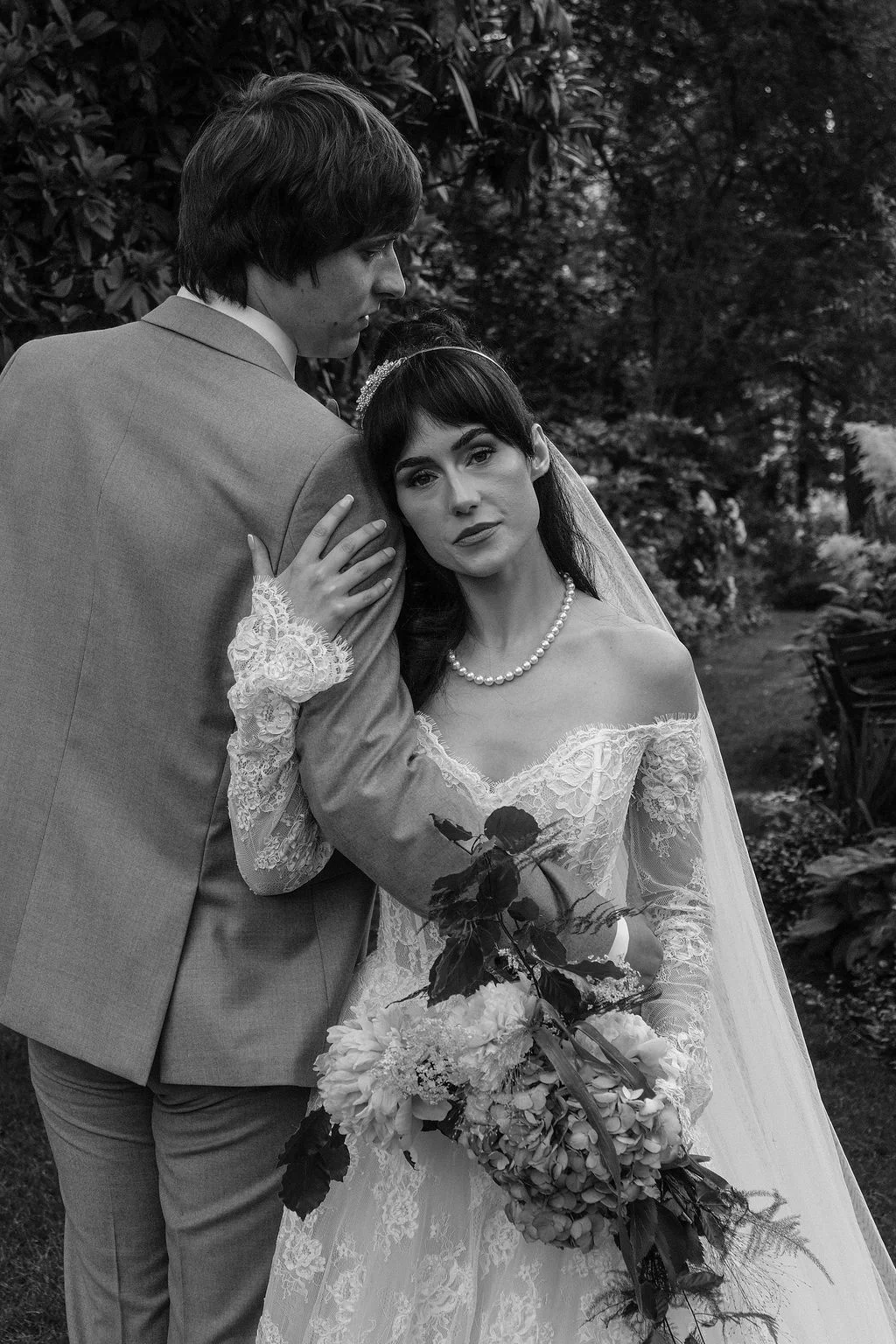 A newlywed couple poses together outdoors in black and white, with the bride resting her head against the groom's shoulder while holding a lush floral bouquet.