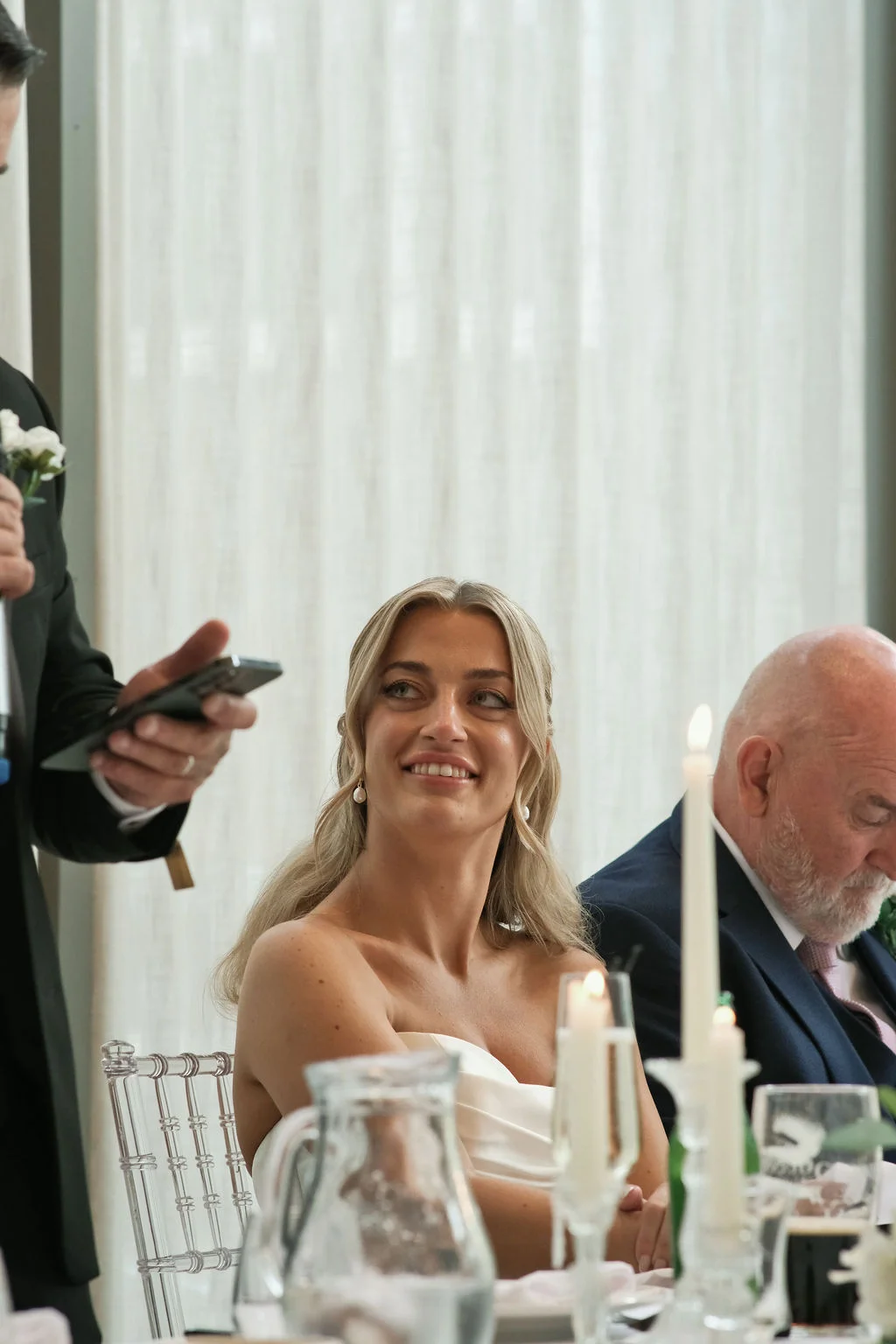 A smiling bride looks up and listens to the wedding speeches during the wedding breakfast, seated at the top table decorated with candles.
