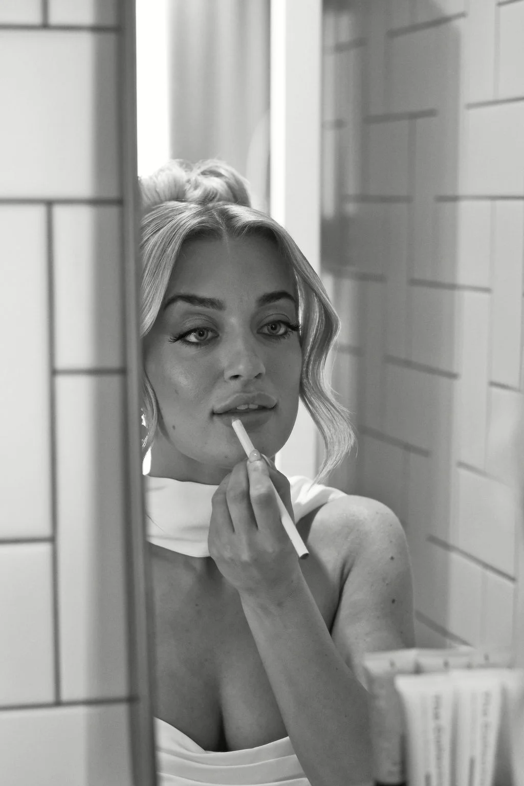 A close-up black and white shot of a bride applying lipstick while looking into a tiled mirror during her morning preparations.
