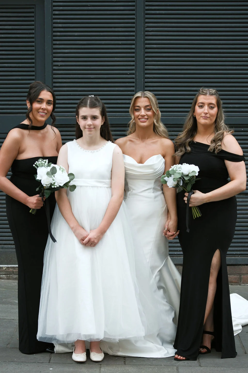 A bride in a white satin strapless gown stands flanked by bridesmaids in black dresses and a young flower girl in white.