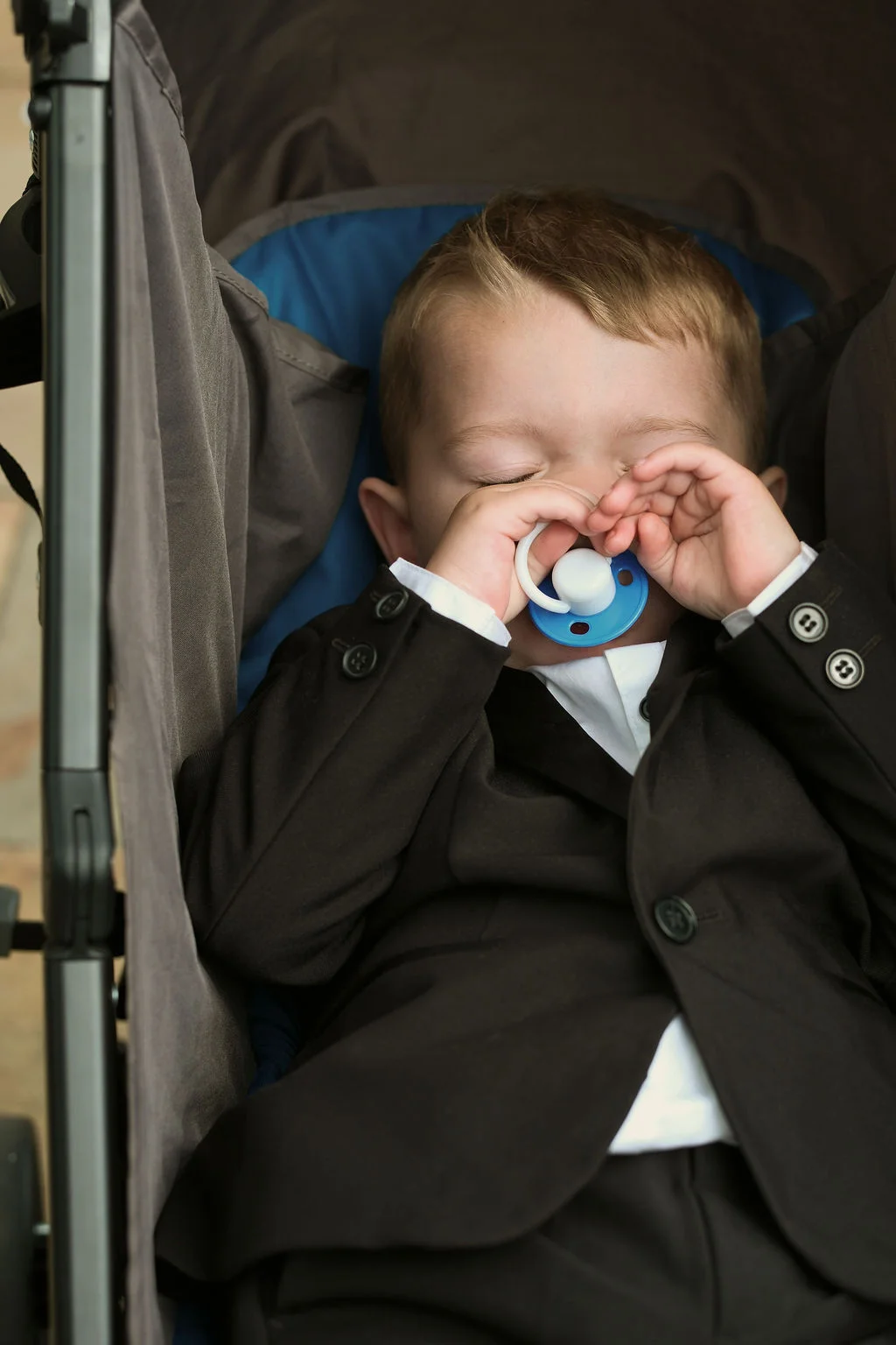 A tiny page boy dressed in a smart black suit and white shirt sleeps peacefully inside a pram with a blue and white dummy.