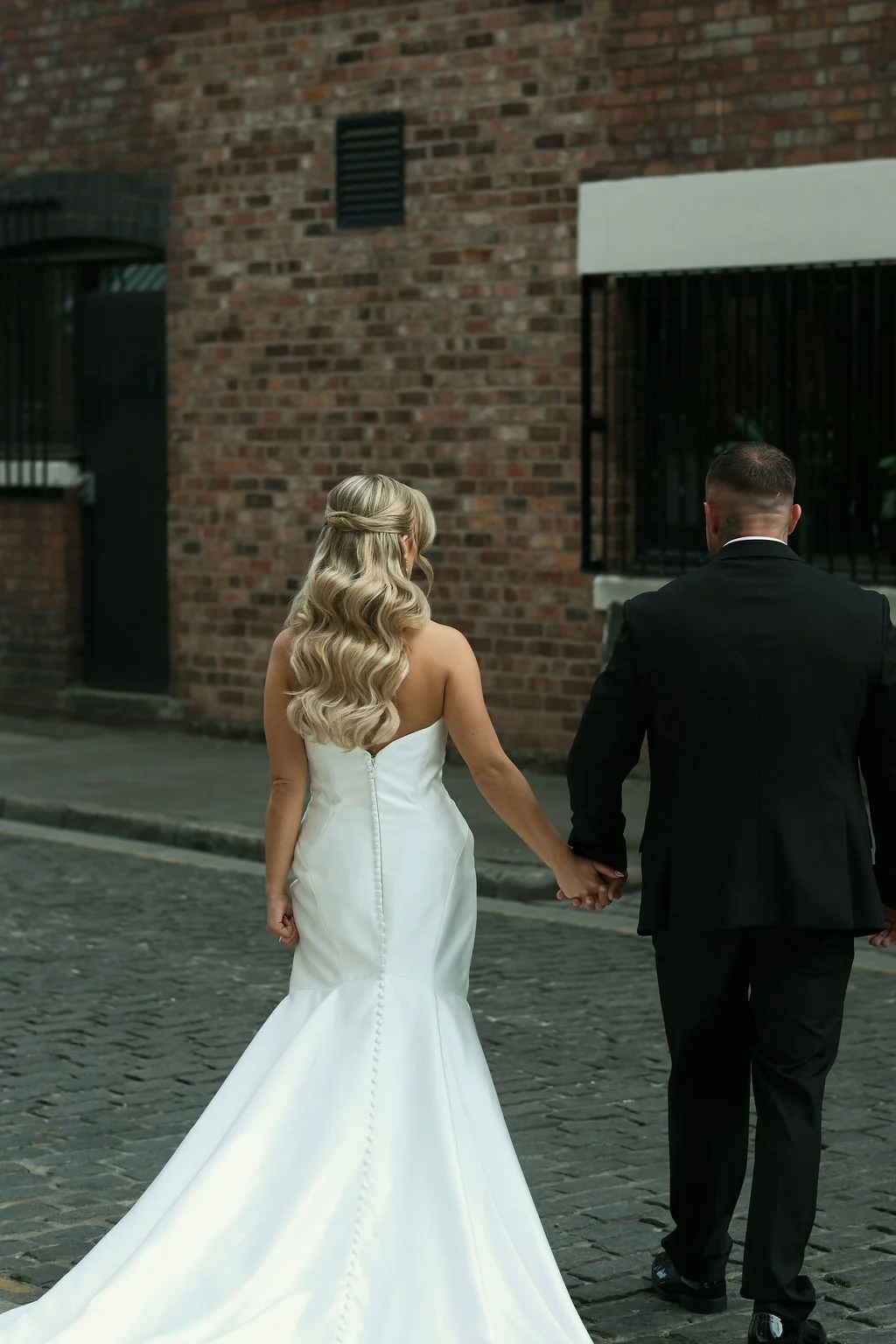 A newlywed couple walks away hand in hand down a cobbled city street lined with exposed brick walls, the bride wearing a strapless satin trumpet gown with a buttoned back and soft flowing waves.