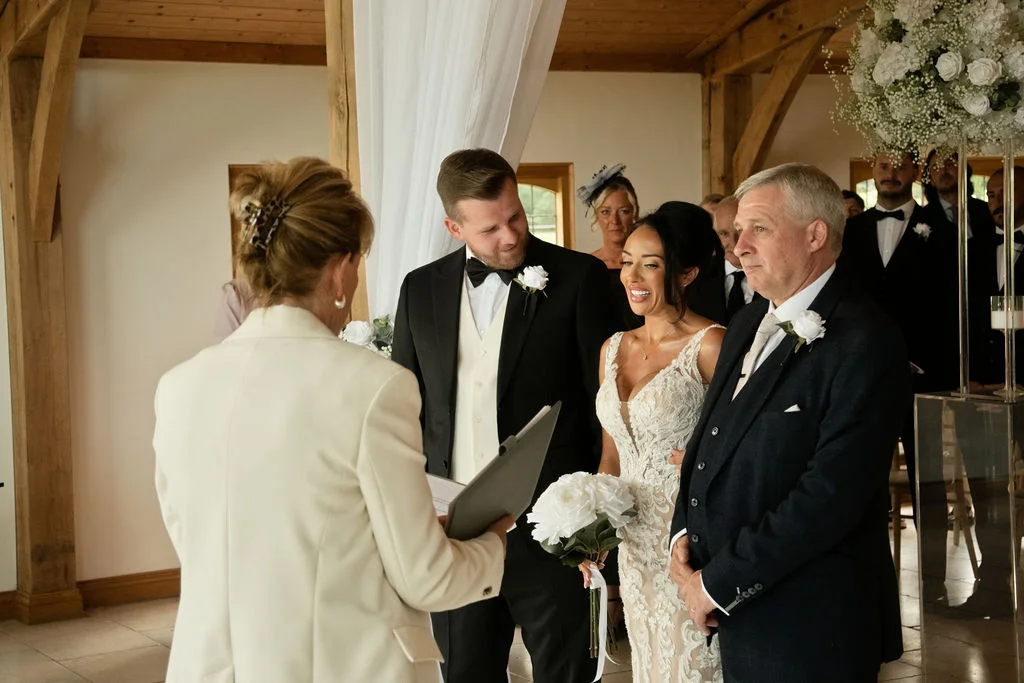 A smiling bride holding a white rose bouquet stands alongside her groom and father during a civil ceremony inside a light-filled oak-framed barn.