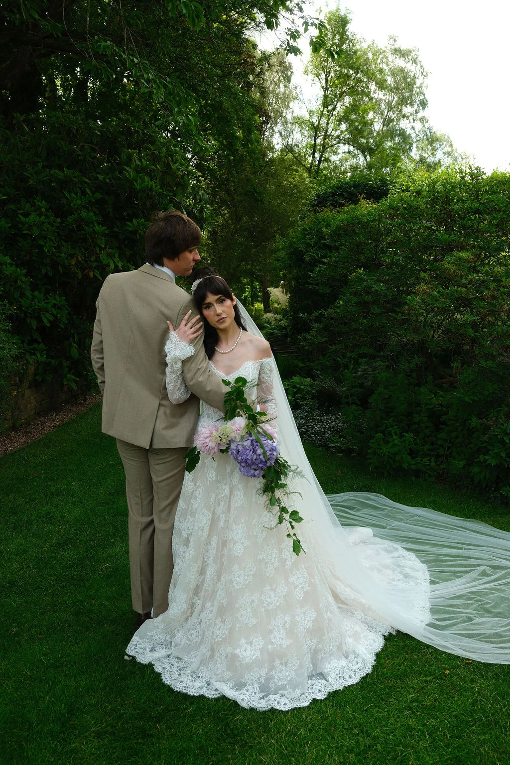 A bride in a lace off-the-shoulder gown holding a cascading bouquet and a groom in a brown suit pose together in a lush garden setting.