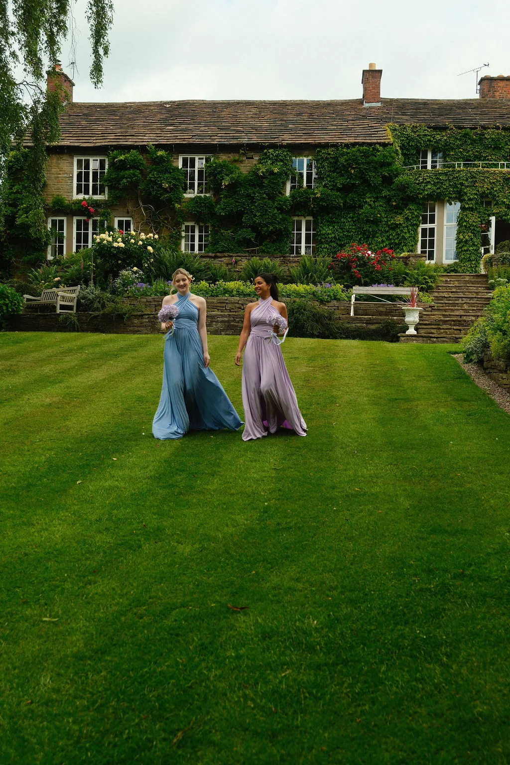 Two smiling bridesmaids in halterneck chiffon dresses walk across a sprawling lawn in front of an ivy-clad country house holding small bouquets.