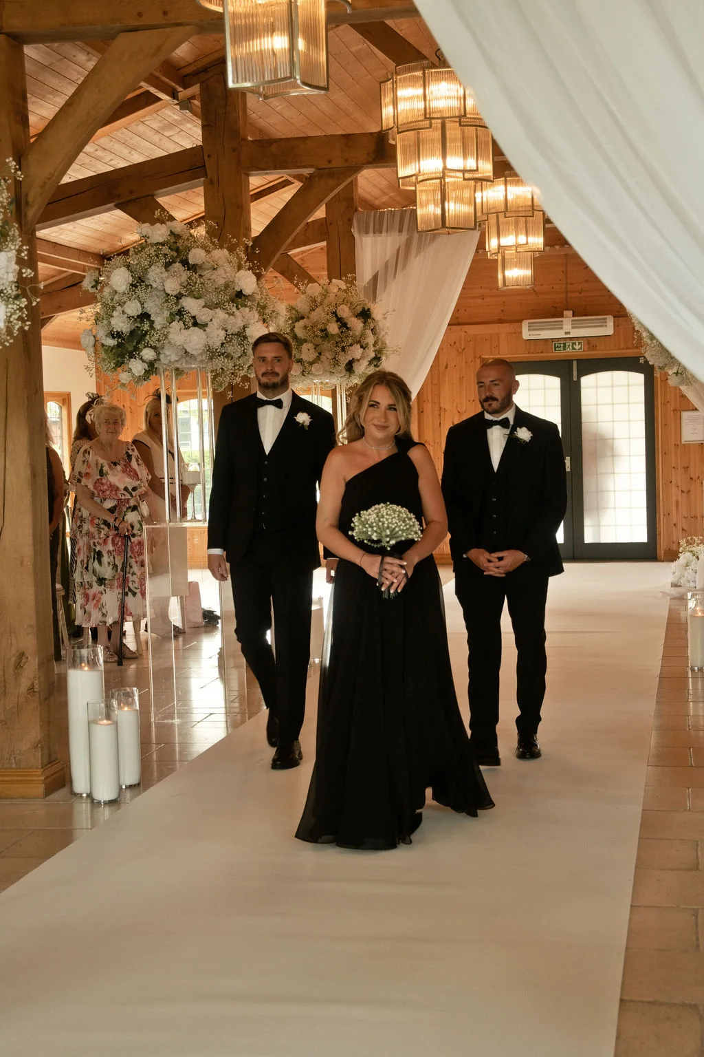 A bride in a striking black one shoulder wedding dress and groom in a classic tuxedo walk down the aisle together inside a rustic oak framed barn.